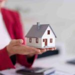 Person in red blazer holding a small white house model, with a calculator nearby.